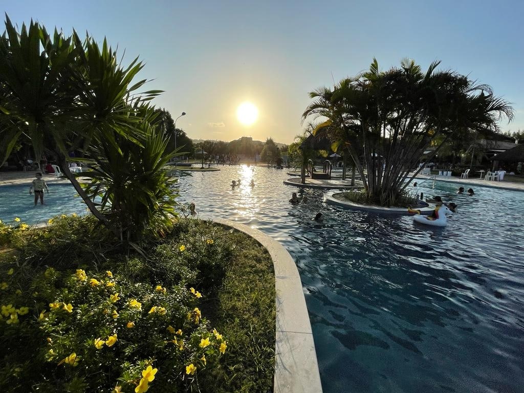 A resort-style pool is surrounded by lush greenery and blooming yellow flowers. The sun sets in the background, casting a warm glow on the water. Guests can be seen enjoying the pool, with several inflatable floats adding to the atmosphere.