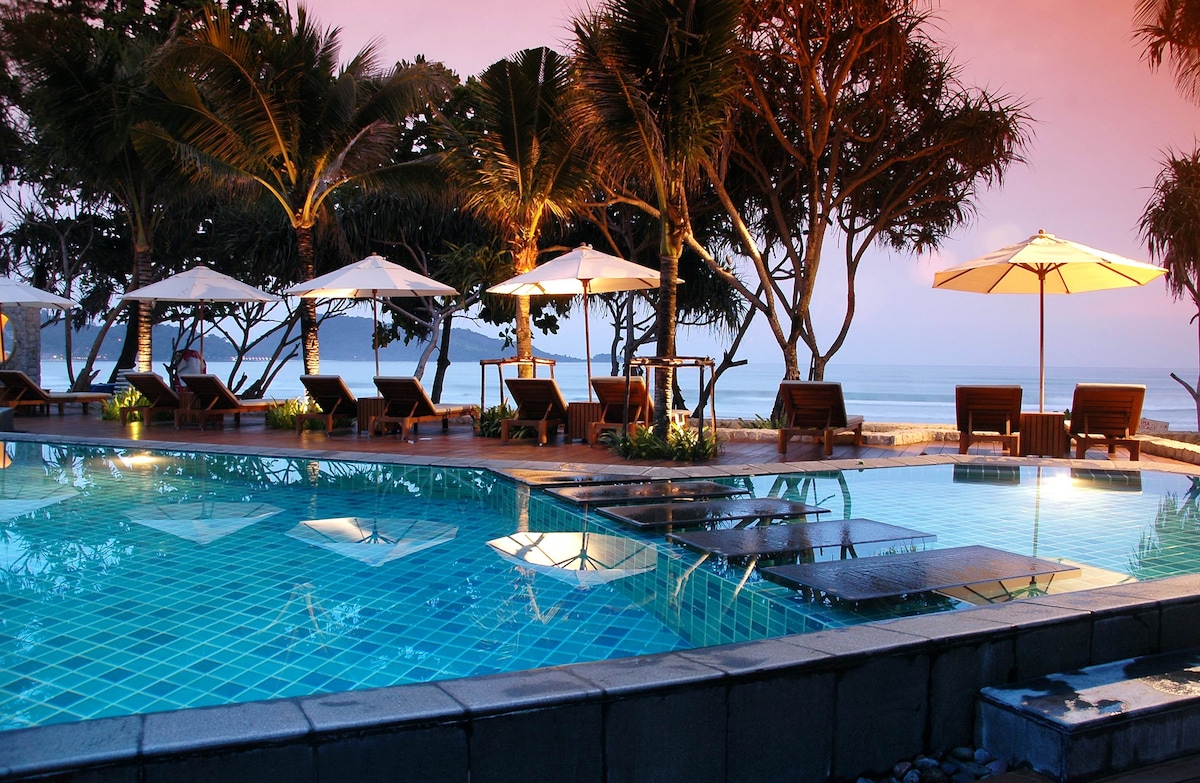 An infinity-edge swimming pool reflects the evening sky, surrounded by palm trees and loungers. Umbrellas provide shade for seating areas, creating a serene setting near the beachfront. Soft lighting highlights the tranquil atmosphere as the water glimmers under the fading sunlight.