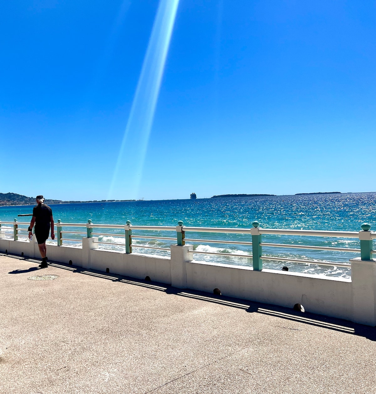 A serene seaside walkway is depicted, showcasing the shimmering turquoise water under a clear blue sky. The sun reflects off the waves, creating a calming atmosphere. A person is seen walking along the path, with distant coastline visible along the horizon.
