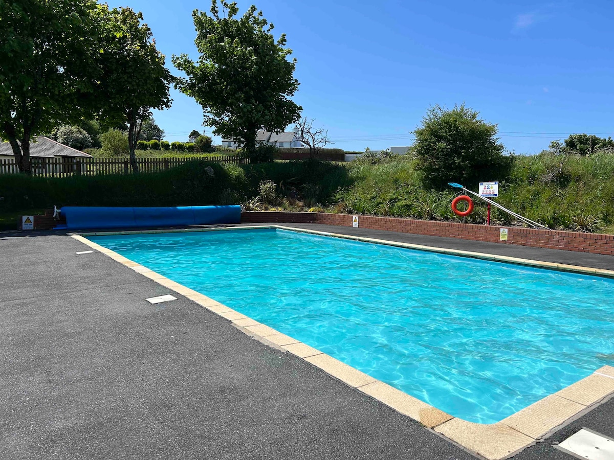 An outdoor swimming pool is captured under clear blue skies, featuring vibrant turquoise water. Surrounding greenery adds a natural touch, while a safety buoy is positioned nearby. The pool area includes a concrete deck with designated lane markings.
