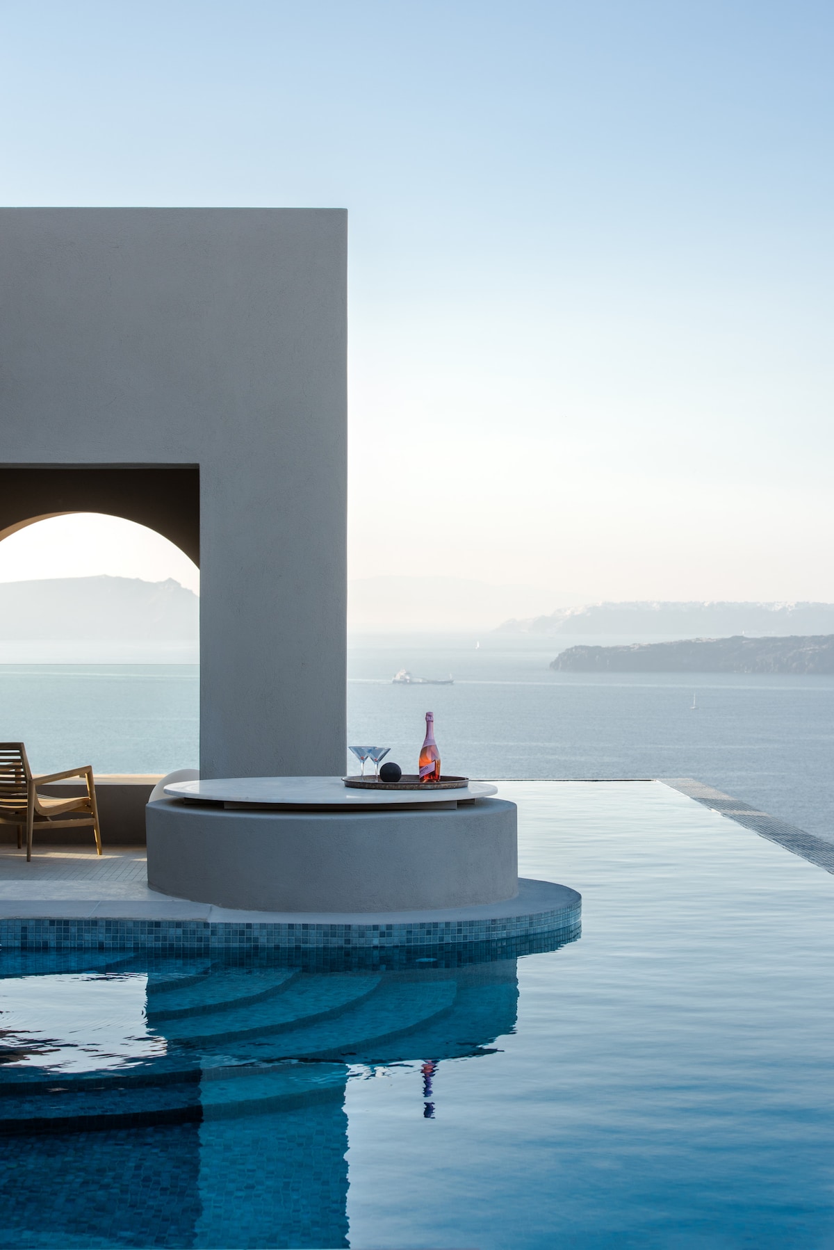 A serene outdoor space features a heated pool with an infinity edge, seamlessly blending with the sea. A round table is set with two glasses and a bottle, while minimalist wooden chairs are positioned nearby, offering stunning views of the horizon and distant hills.