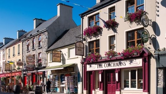A charming streetscape features the Coachman's establishment, showcasing an inviting façade with hanging planters adorned with vibrant flowers. Nearby shops and the local pub contribute to the lively atmosphere, framed against a clear blue sky.