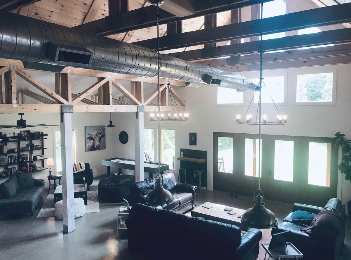 The spacious common room features high wooden ceilings with exposed beams and modern metal ductwork. Comfortable seating arrangements, including leather sofas, are positioned around a central coffee table. Large windows allow natural light to fill the space, creating an inviting environment that encourages social interaction.