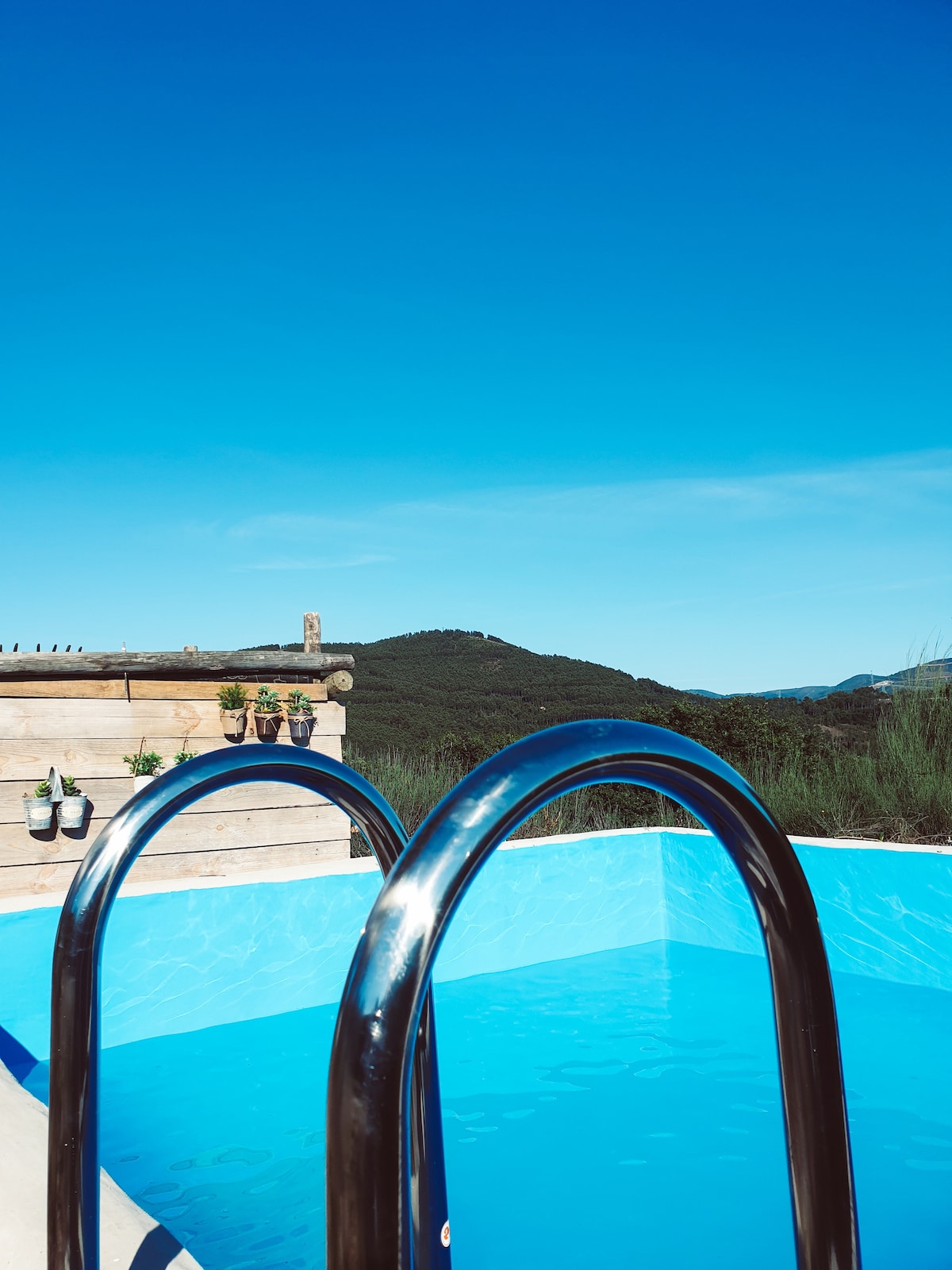 A clear blue swimming pool is framed by a modern wooden deck. Two silver handrails provide access to the water, reflecting the bright blue sky. Lush green hills are visible in the background, adding to the natural setting.