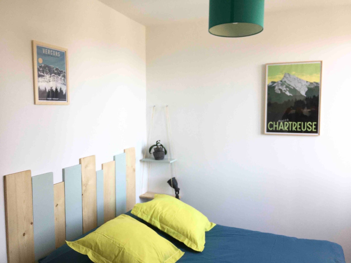 A comfortable sleeping area is presented with a wooden headboard and bright yellow pillows atop a spacious bed. A framed mountain-themed artwork adorns the wall, while a shelf holds a decorative kettle. Natural light filters in, enhancing the serene atmosphere.
