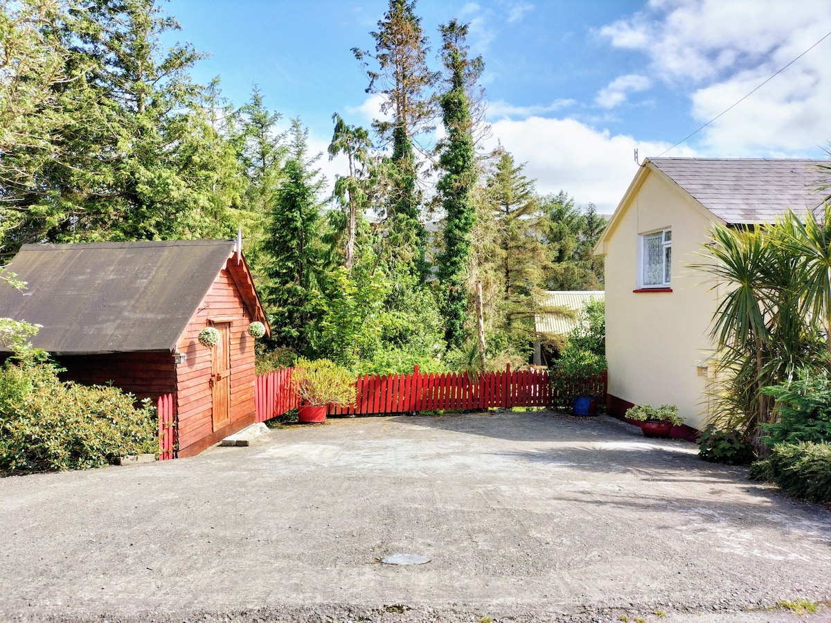 A stone driveway leads to the cottage, framed by an array of greenery and trees. A red picket fence encloses a well-maintained garden area, with potted plants adding a touch of color. The two-story cottage is visible on the right, contrasted against the natural surroundings.
