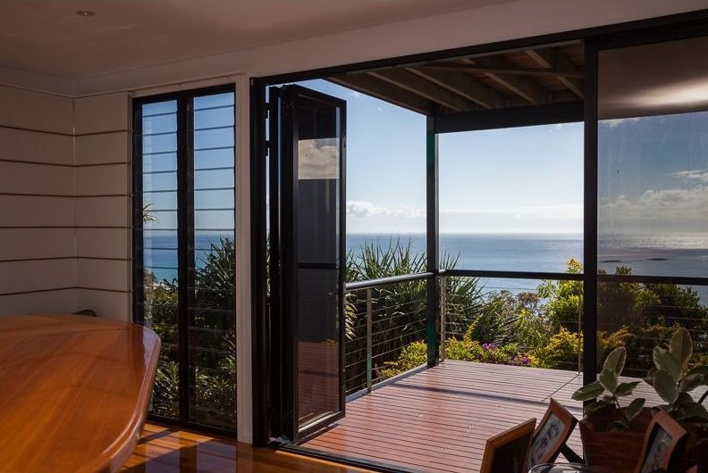 Expansive sliding doors open to reveal a scenic view of the Coral Sea. The outdoor deck is framed by greenery and features wooden flooring. An inviting dining area with a polished wooden table is visible in the foreground, enhancing the connection between indoor and outdoor spaces.