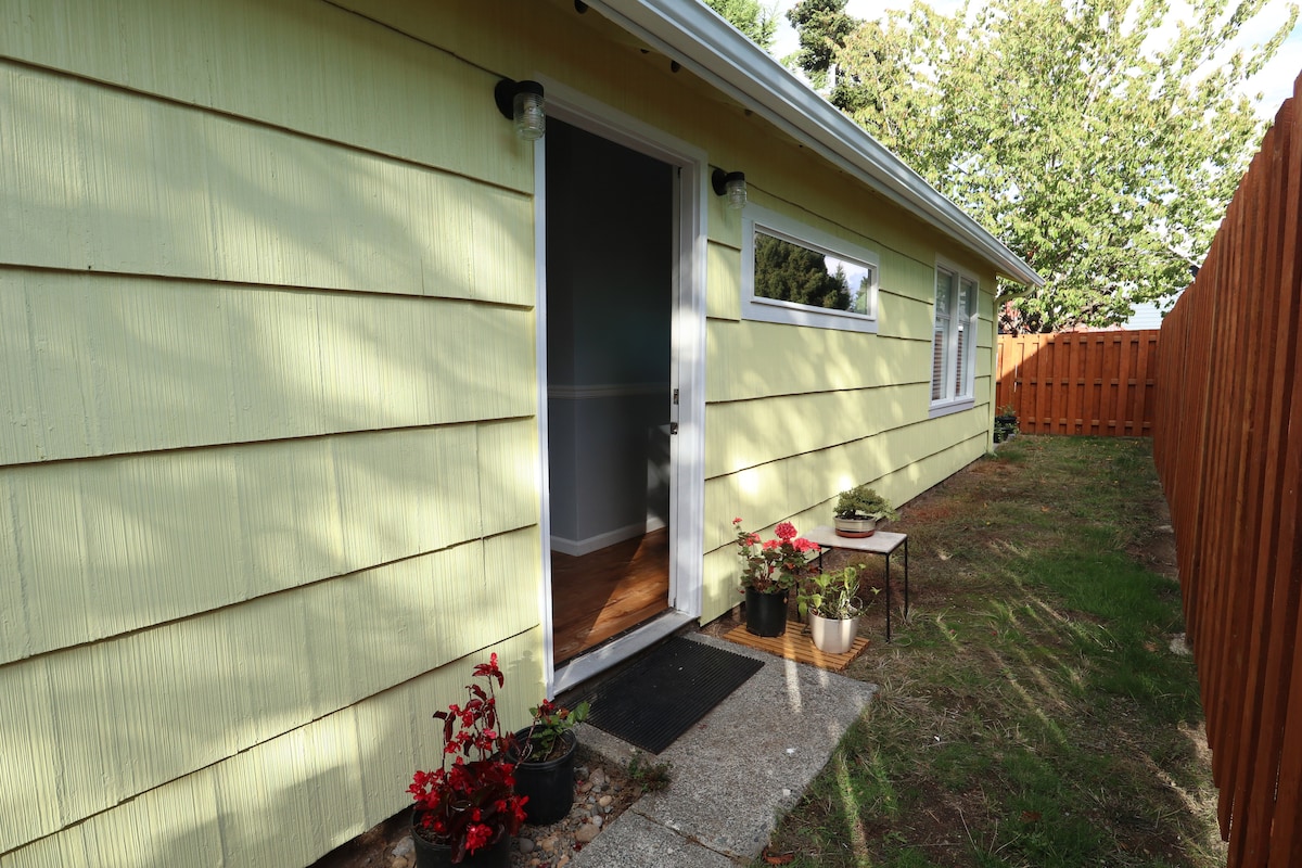 A side view of a single-story yellow bungalow is shown, featuring a welcoming entry with a door and a small concrete step. Potted plants with vibrant red flowers are placed by the entrance, while a grassy area extends toward a wooden fence set in the background.