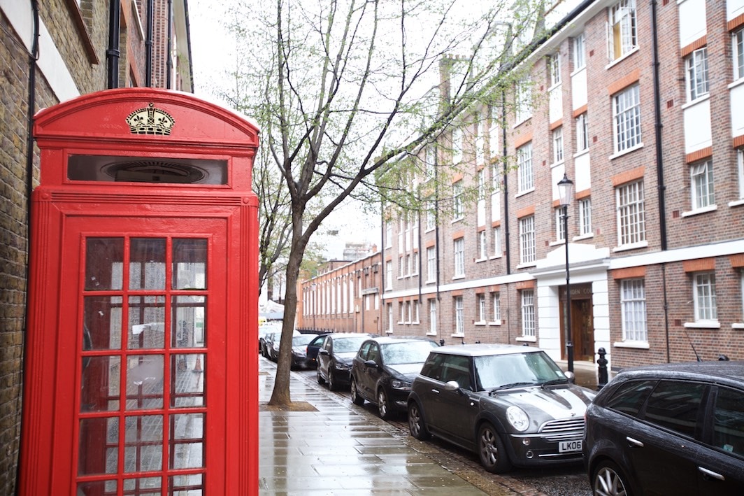 A traditional red phone booth stands on a rain-soaked street, accompanied by trees with budding leaves. The adjacent row of brick buildings showcases large windows, while various parked cars line the curb, providing a glimpse of the urban environment.