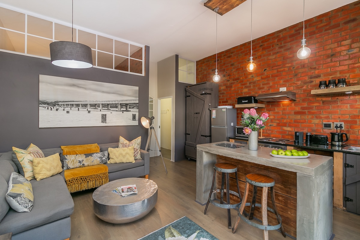 A modern living space features a comfortable grey sectional sofa adorned with patterned cushions. A rustic wooden dining table with stools complements the brick accent wall. A flower vase adds a touch of color, while a magazine lies on the table beneath a softly lit pendant light.