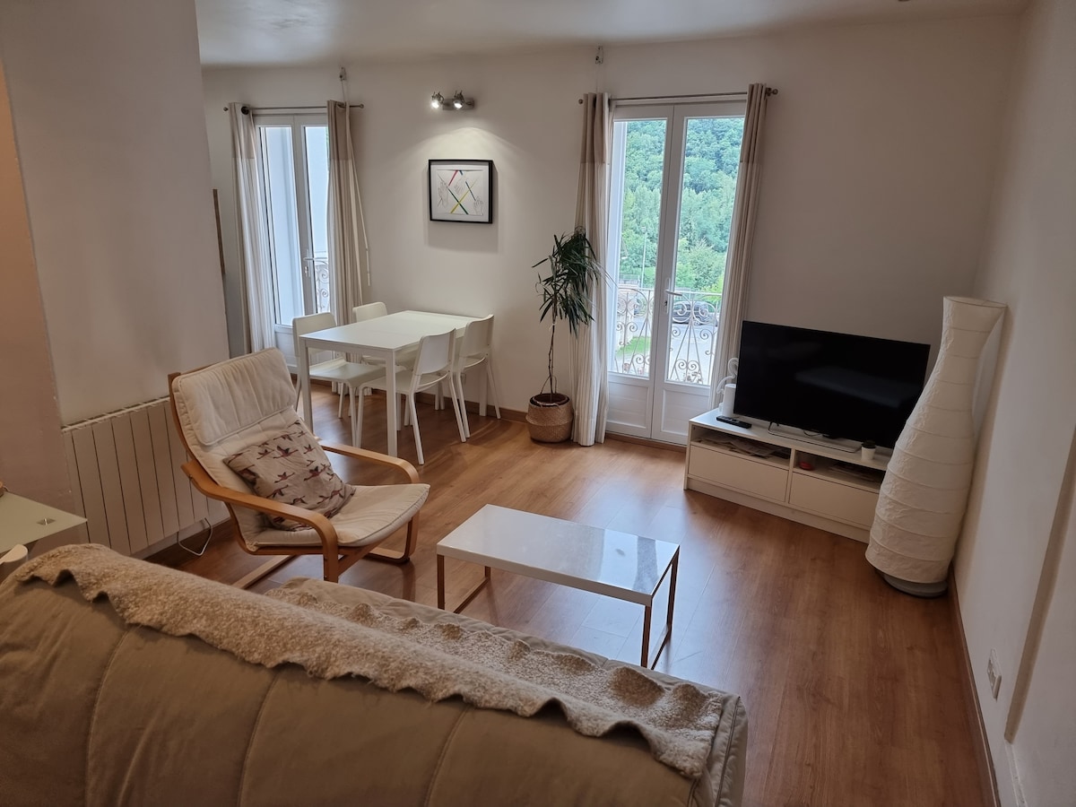 A well-lit living area features a comfortable armchair and a sleek white coffee table. A dining table with four chairs is positioned near large windows, offering a view of greenery outside. A television is placed on a low console against the wall, adding functionality to the space.