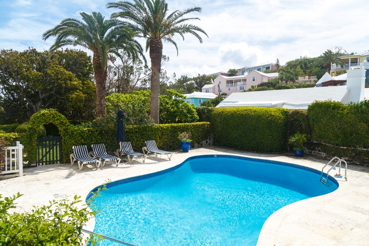 A serene pool area is surrounded by lush greenery and palm trees. Relaxing lounge chairs are positioned near the water, inviting guests to unwind in the tranquil setting. A blue pool sparkles under the sunlight, offering a refreshing retreat.