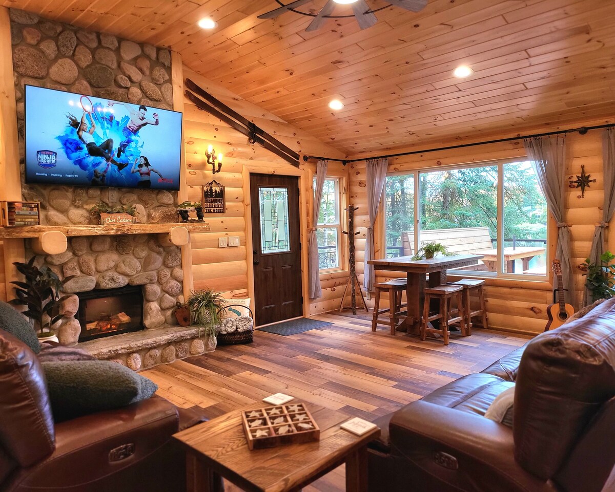 A warm living space features wood-paneled walls and a stone fireplace. A large television is mounted above the hearth. Natural light fills the room through expansive windows, and a cozy dining table is positioned near the entrance, complemented by comfortable leather seating.