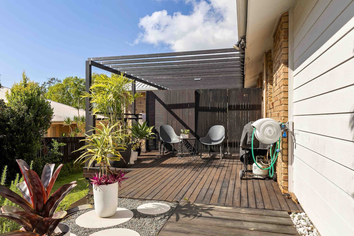 A private deck is featured, surrounded by greenery and potted plants. Comfortable seating options are provided, with a shaded area created by a wooden pergola above. A hose and outdoor accent items are also visible, contributing to a cozy outdoor space.