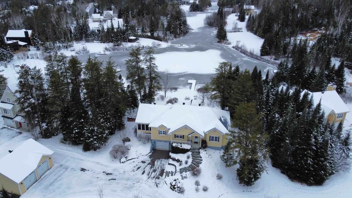 An aerial view captures the LakeHouse nestled in a snowy landscape. The property is surrounded by trees and features a private beach area along the iced-over Lake Lepage. The snow-covered roof displays a welcoming structure amid the serene winter setting.