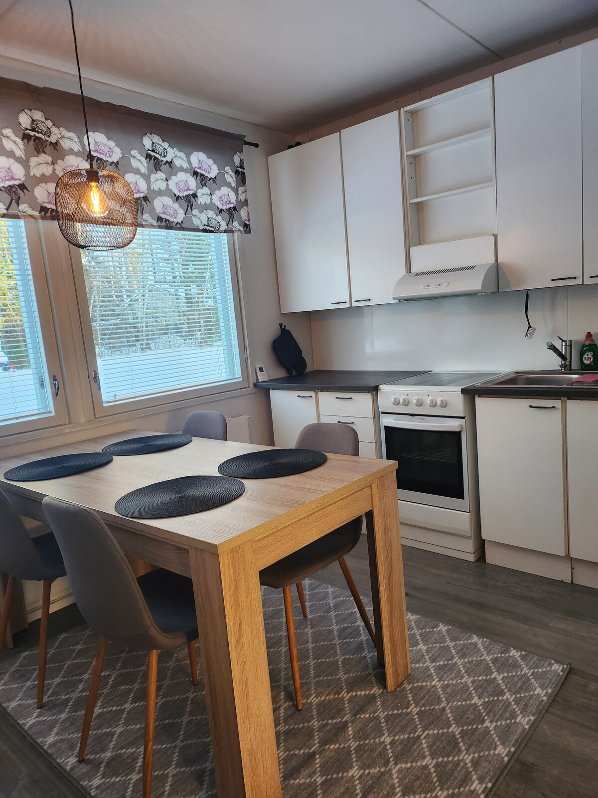 A bright kitchen area is equipped with modern appliances, including an oven and stovetop. A wooden dining table, accompanied by four gray chairs, is positioned under a stylish light fixture. Natural light filters through the windows, adorned with floral curtains.