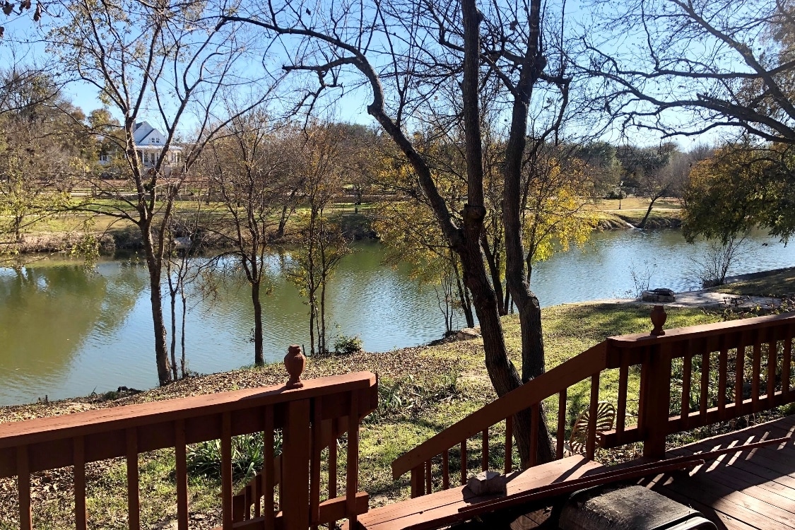 A view from a wooden deck overlooks the Paluxy River, flanked by trees with autumn foliage. Gentle ripples are seen on the water's surface, while steps lead down from the deck to the riverbank, providing an inviting access point to the natural surroundings.