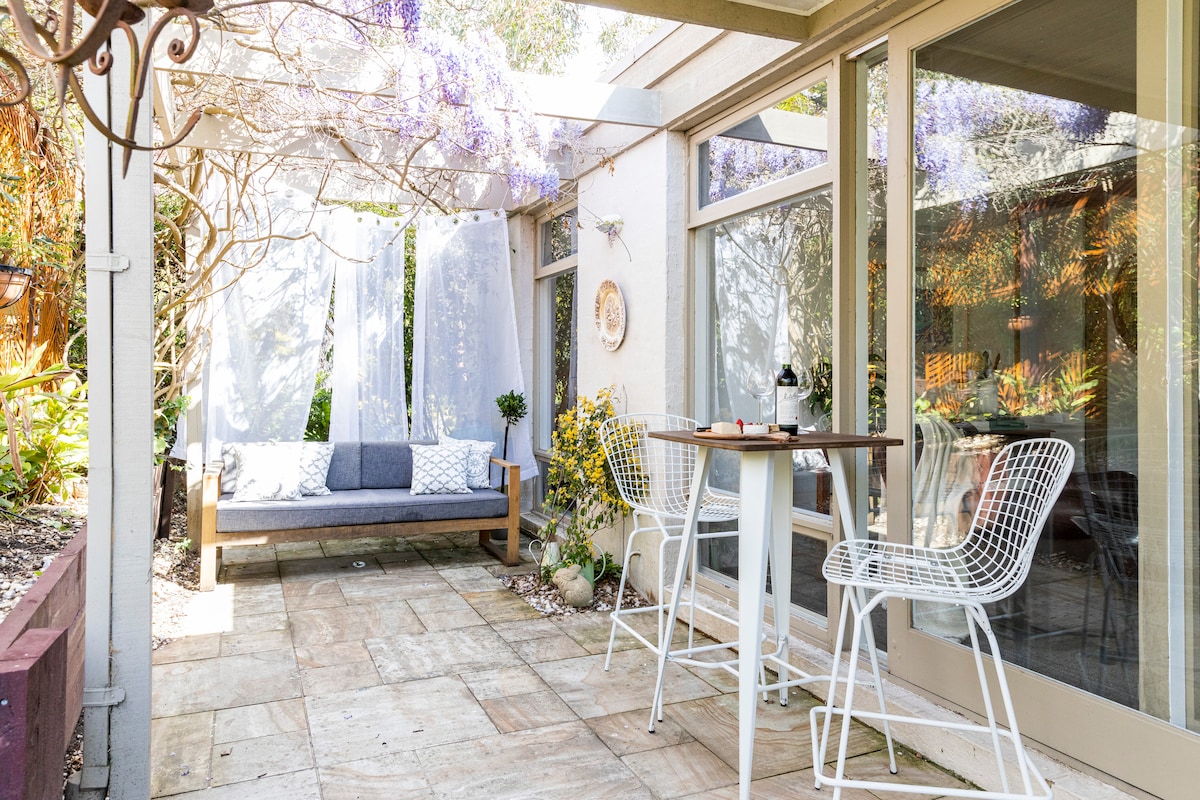 An outdoor terrace area is depicted, featuring a comfortable day bed with cushions and white decor. A small table with two chairs is positioned nearby, surrounded by greenery. Soft purple flowers and natural light create a serene outdoor environment.