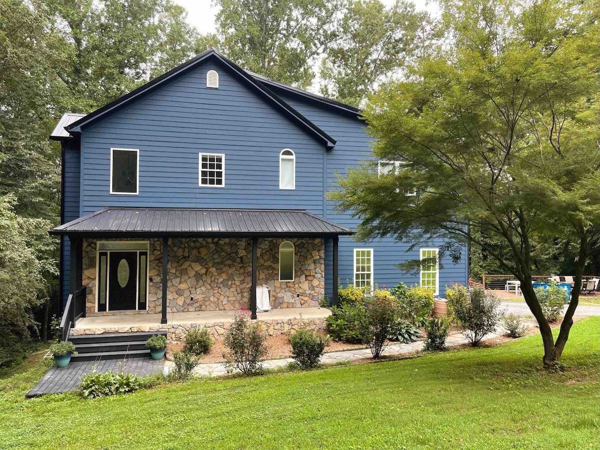 A two-story lakefront home is showcased, featuring a mix of blue siding and stone accents. A covered porch with an oval entrance door is visible, framed by greenery and well-maintained landscaping. Ample natural light enters through various windows, enhancing the welcoming facade.