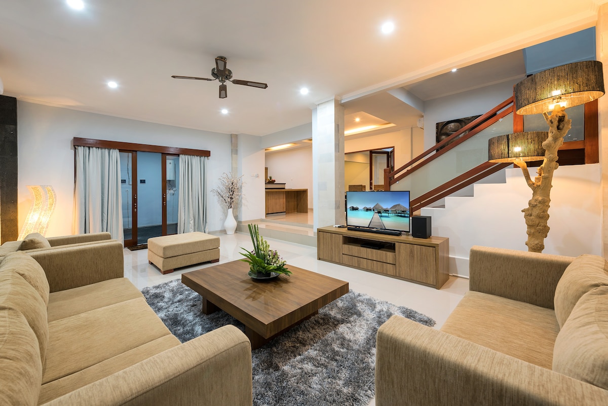 A spacious living area features light-colored sofas arranged around a wooden coffee table, with a flat-screen television positioned on a console. Natural light fills the room, highlighting the clean lines and modern design. A ceiling fan is visible, providing comfort in the inviting space.