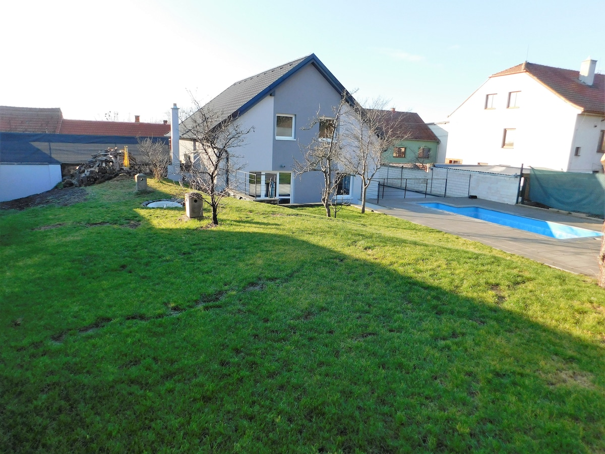 A private house is set within a spacious green yard, featuring several mature trees and a paved area. An outdoor pool is visible, enclosed by a fence. Nearby, additional houses can be seen, with clear sunny skies overhead.