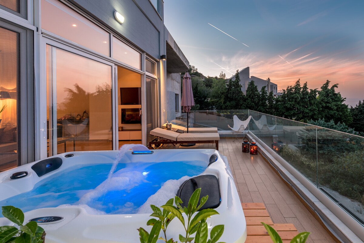 A private balcony features a hot tub, surrounded by greenery and wooden decking. Glass railings provide unobstructed views of the coastline, while evening light casts a soft glow. Comfortable lounge chairs are positioned nearby, creating a serene outdoor space for relaxation.