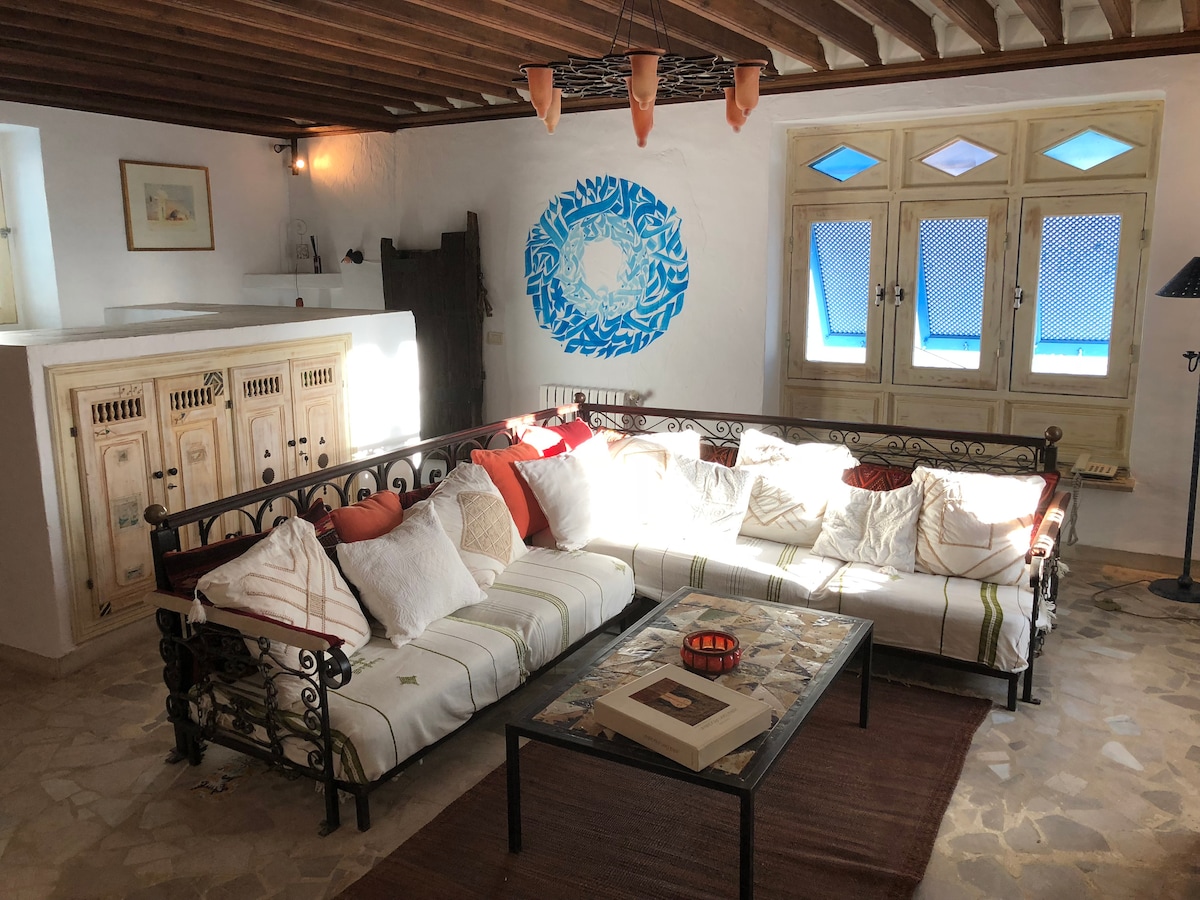 A spacious living area features a large, inviting sofa adorned with assorted cushions. A coffee table sits at the center, surrounded by natural light streaming through three decorative blue windows. The walls are illuminated by soft lighting, enhancing the room's relaxed ambiance.