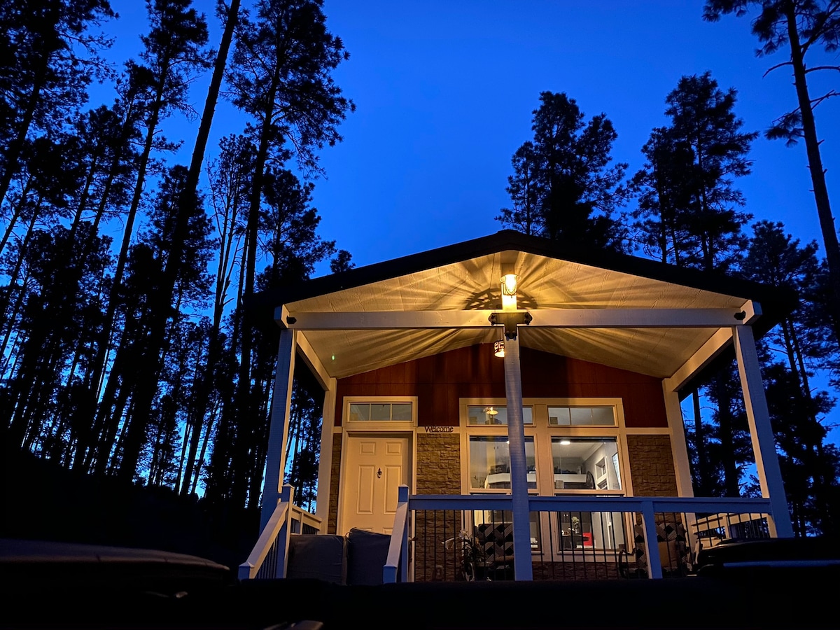 The cabin is viewed from below against a deep blue evening sky, surrounded by tall pine trees. Soft lighting highlights the porch and entrance, inviting guests into the cozy interior. Large windows reveal hints of the stylish decor inside.