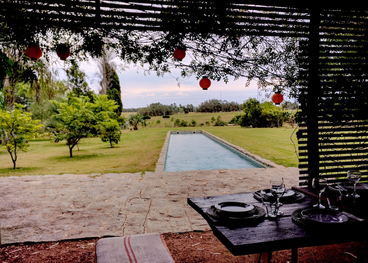 An outdoor dining area is shaded by a wooden trellis, with a view of a swimming pool surrounded by lush greenery. The table is set for a meal, with plates and glassware arranged, while soft lighting fixtures hang overhead, enhancing the serene atmosphere.