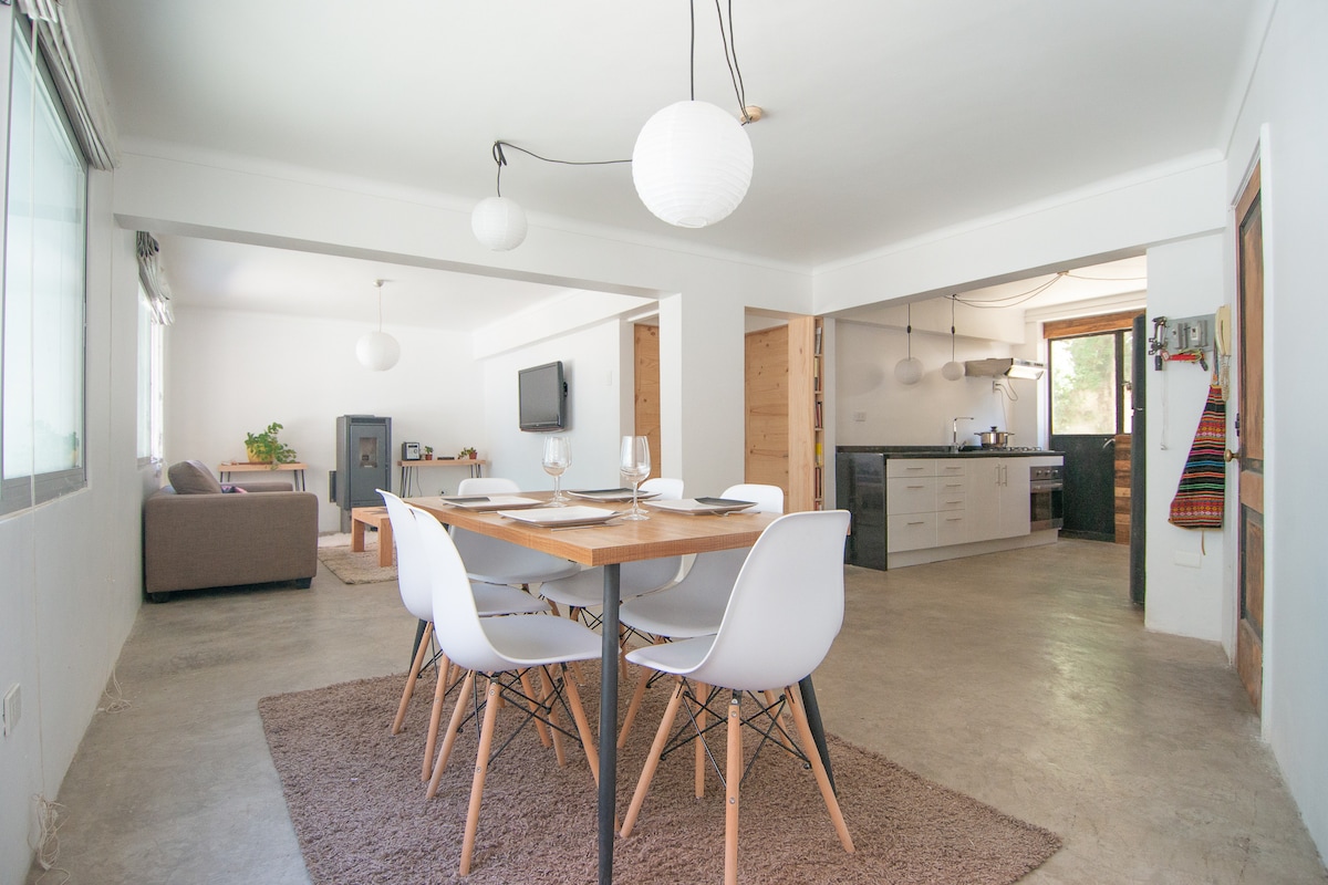 A dining area features a wooden table surrounded by white chairs, with a plush rug underneath. Natural light enters through large windows, illuminating the space. A cozy living area with a sofa and a wall-mounted TV is visible in the background, alongside an open kitchen.