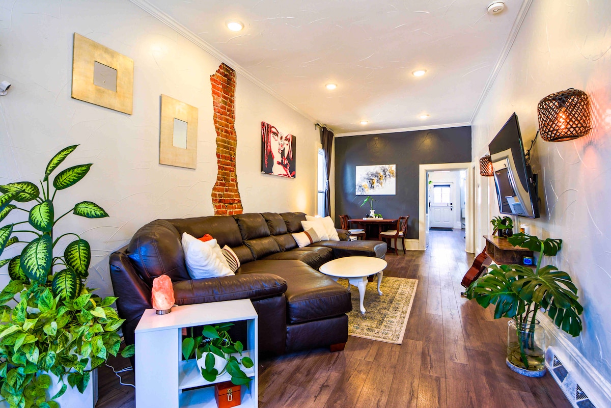 A spacious living room is highlighted by a large, dark sectional sofa and a round coffee table. A muted decorative rug sits on the hardwood floor. Plants and a salt lamp add a touch of greenery. Wall art and accent lighting create a modern ambiance.