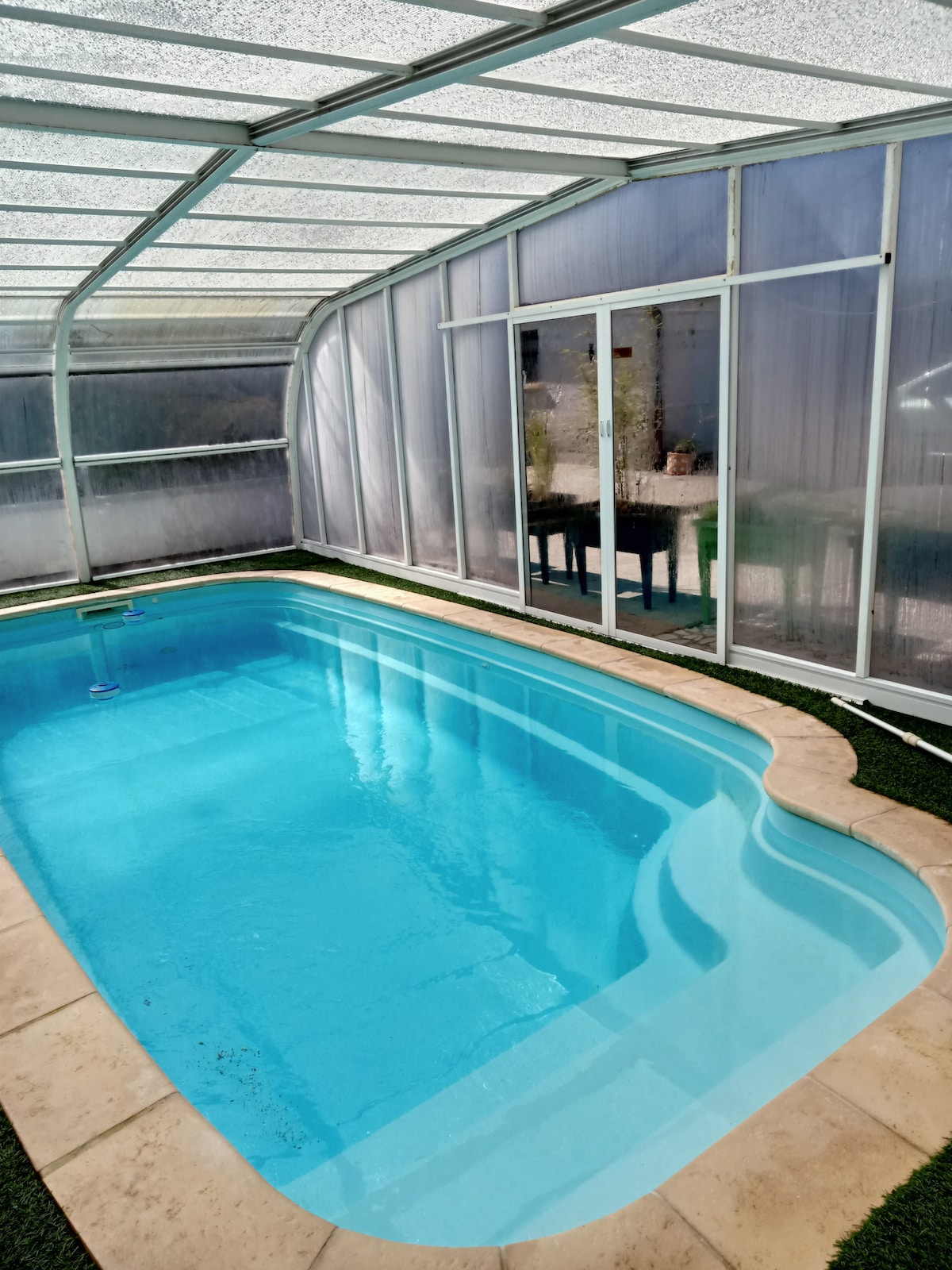 An indoor pool is positioned beneath a transparent roof, surrounded by greenery and light-colored tiles. The water appears clear and inviting, reflecting natural light from the windows, while a table is visible in the background, creating a relaxed atmosphere.