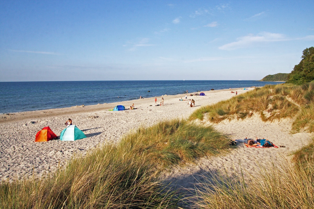 A sandy beach stretches along the shoreline, bordered by gentle dunes of tall grass. Colorful beach tents dot the landscape, while visitors enjoy the sun and surf. The water is calm, reflecting a clear blue sky above, creating a serene coastal scene.