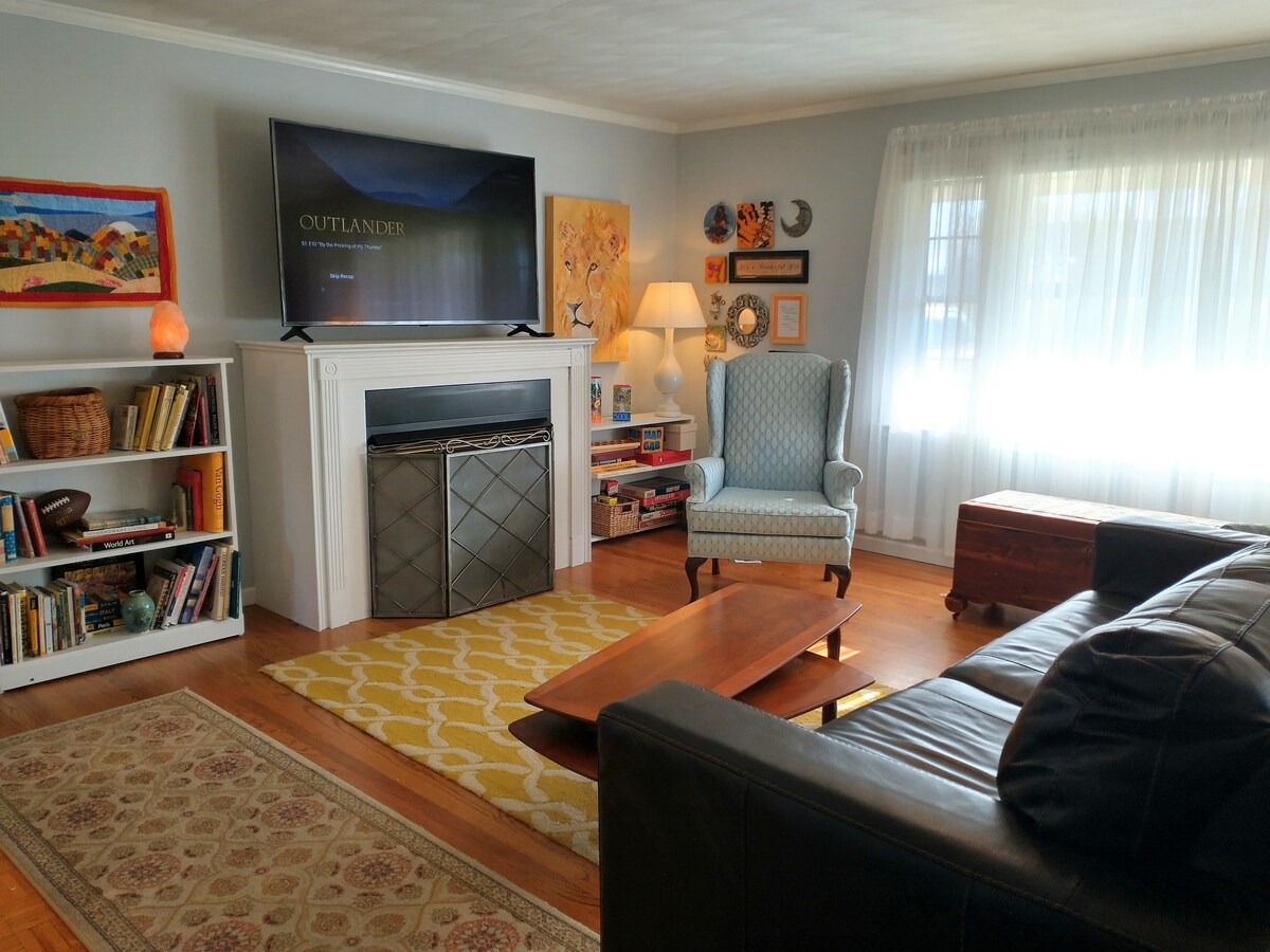 The inviting living room features a large flat-screen television mounted above a decorative fireplace. A comfortable leather sofa faces a wooden coffee table, while a patterned area rug adds warmth. Natural light flows in through sheer curtains, complemented by curated wall art.