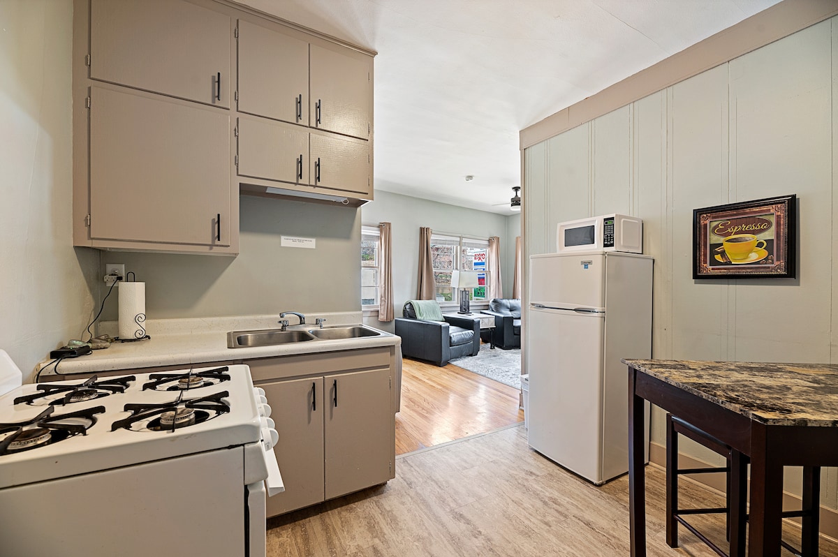 A functional kitchen area features light cabinetry and a compact, gas stove. A refrigerator is positioned adjacent to the countertop, with a small table set nearby. The space is well-lit, revealing wood flooring and textures from the adjoining living area.