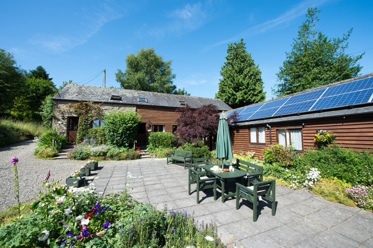 The exterior of Weaver Cottage is shown, featuring a shared stone patio area surrounded by colorful flowers and greenery. Solar panels are positioned on the roof, and several green outdoor chairs and tables are arranged for guest use. The cottage's entrance is visible, marked by a welcoming entrance surrounded by plants.