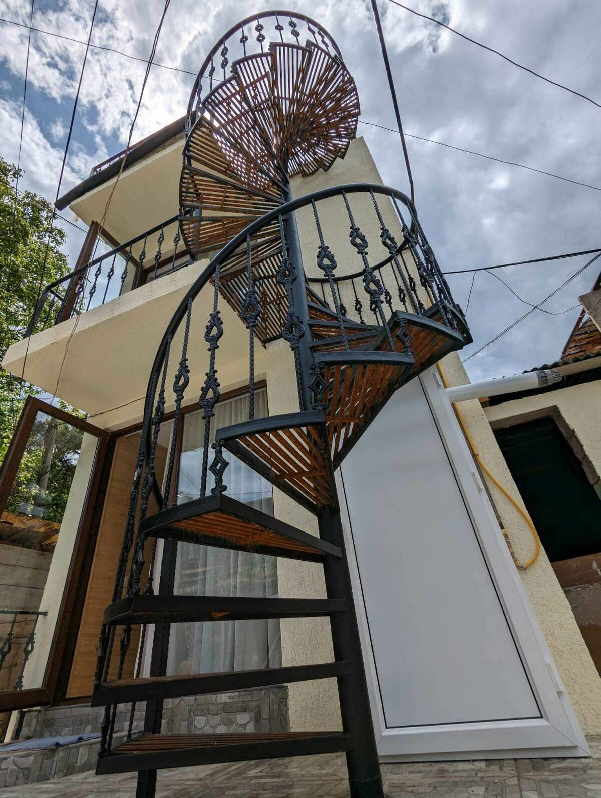 A circular metal spiral staircase is observed, featuring intricately designed railings and wooden steps. The structure ascends alongside a building with a light exterior and large windows, merging gracefully with the surrounding greenery.