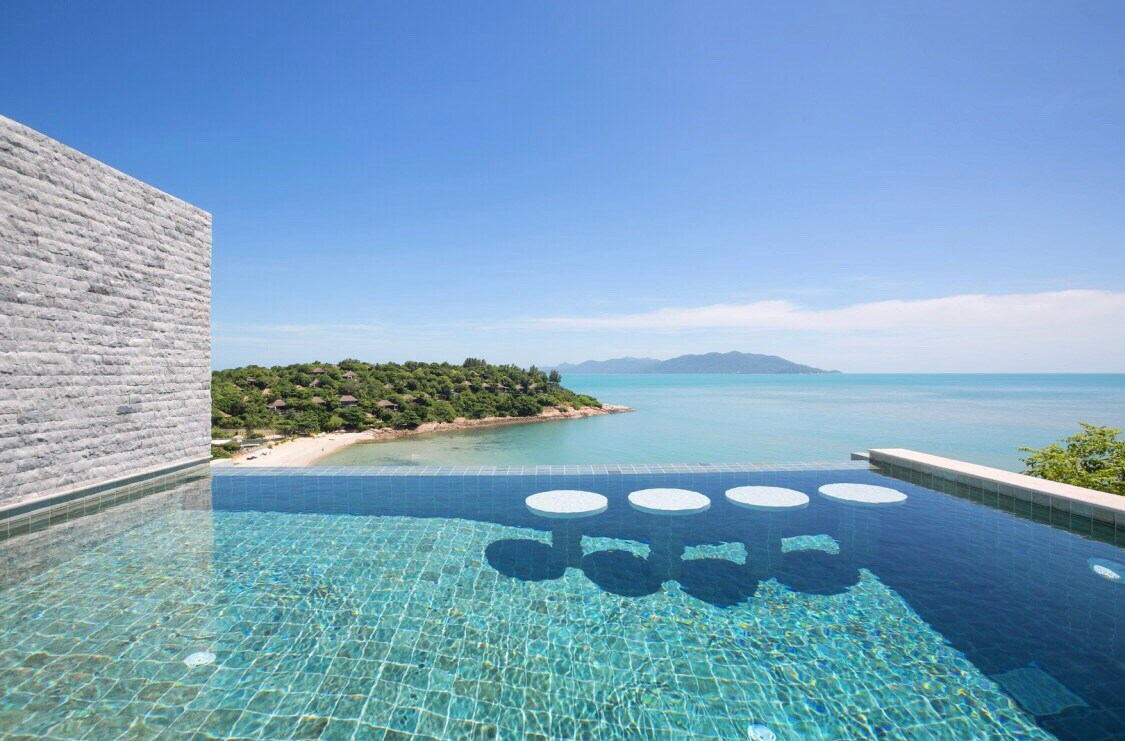 A private infinity pool is shown, merging with the vibrant blue ocean. The pool features circular seating for relaxation. Lush greenery and a sandy beach outline the picturesque landscape, while distant mountains are visible along the horizon under a clear blue sky.
