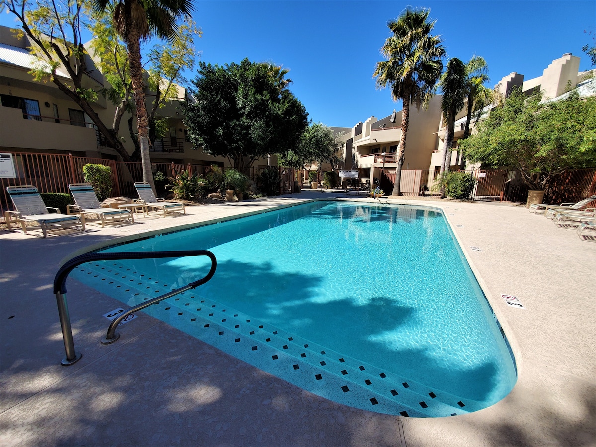 A heated swimming pool is set within a landscaped area, surrounded by palm trees and shrubbery. Lounge chairs are arranged along the pool's edge, while the pool's clear blue water reflects the sunlight on a clear day.