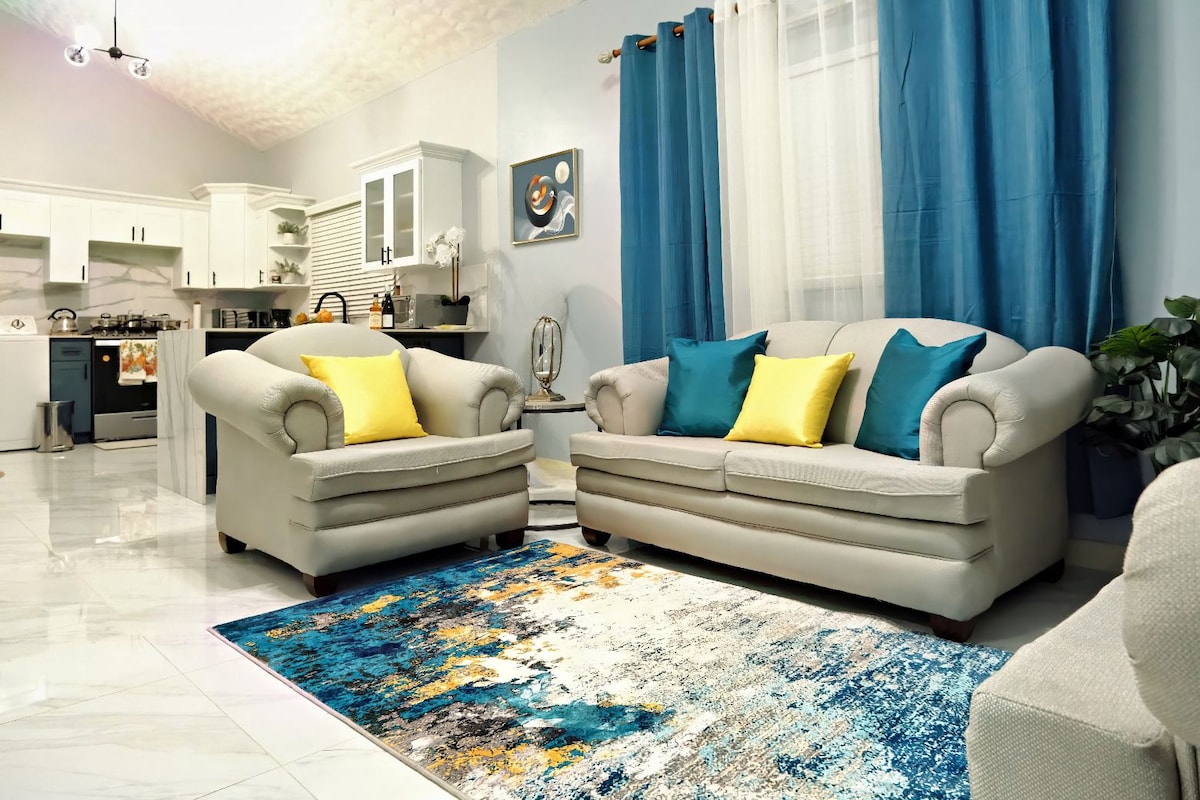 A comfortable living area features two light-colored sofas accented with yellow cushions, set against a backdrop of pale blue walls. A colorful area rug covers the polished tile floor, and large windows are dressed with flowing blue curtains that allow natural light to fill the space.