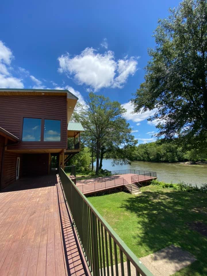 A spacious deck extends along the riverfront, complemented by a grass area and a staircase leading to the water. The lodge features large windows, reflecting the surrounding trees and blue sky, while the serene river flows gently nearby.