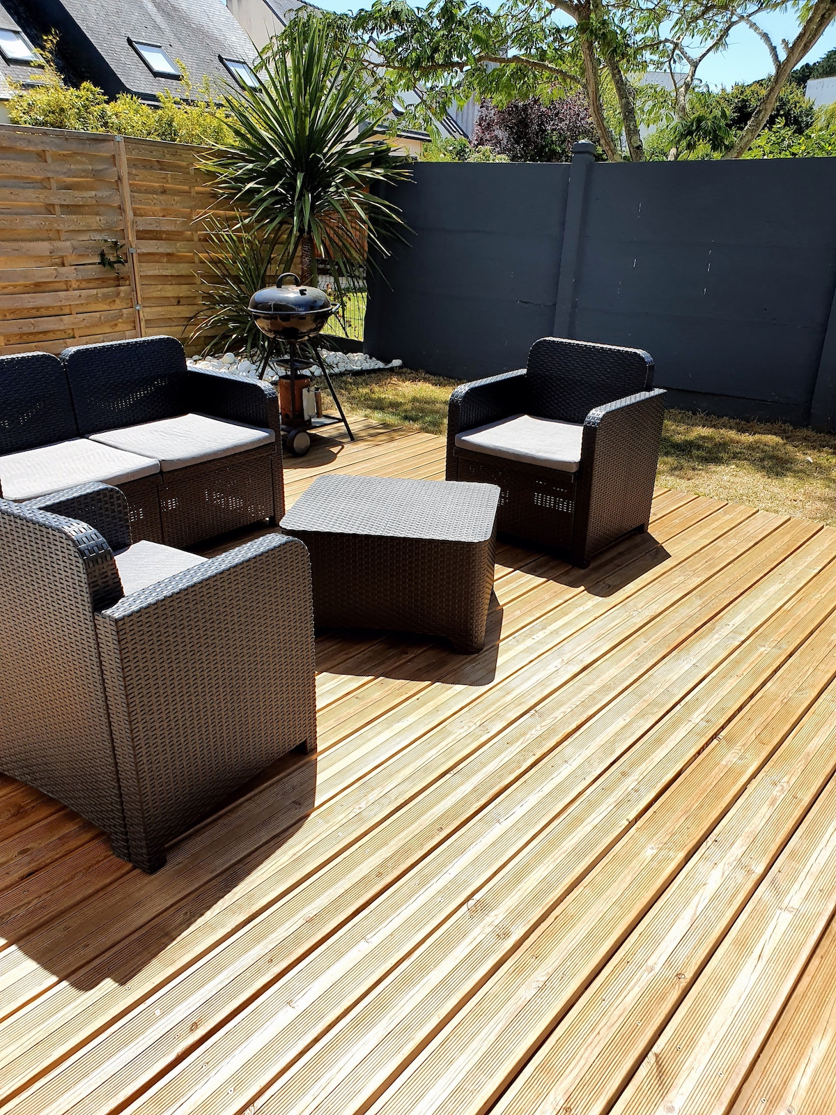 An outdoor terrace features wooden decking with a seating arrangement consisting of a table and four wicker chairs. A small grill is positioned nearby under a green leafy plant beside a gray fence, creating a welcoming space for relaxation.