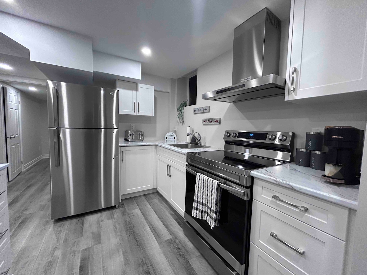 The kitchen features modern stainless steel appliances, including a refrigerator and an oven. White cabinetry is complemented by a marble countertop. Ample workspace is provided, with a toaster and a kettle visible. Soft illumination highlights the clean, organized layout of the kitchen.