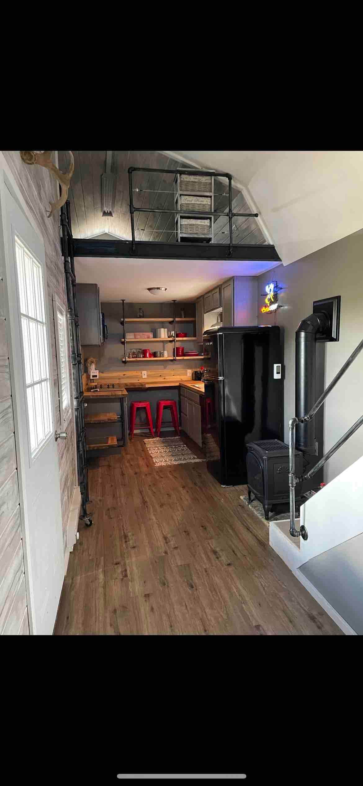 An open kitchen area is highlighted by modern cabinetry and stainless steel appliances. Red stools are positioned at a small counter, complementing the warm wood floors. A staircase leads to an upper level, enhancing the space's vertical appeal.
