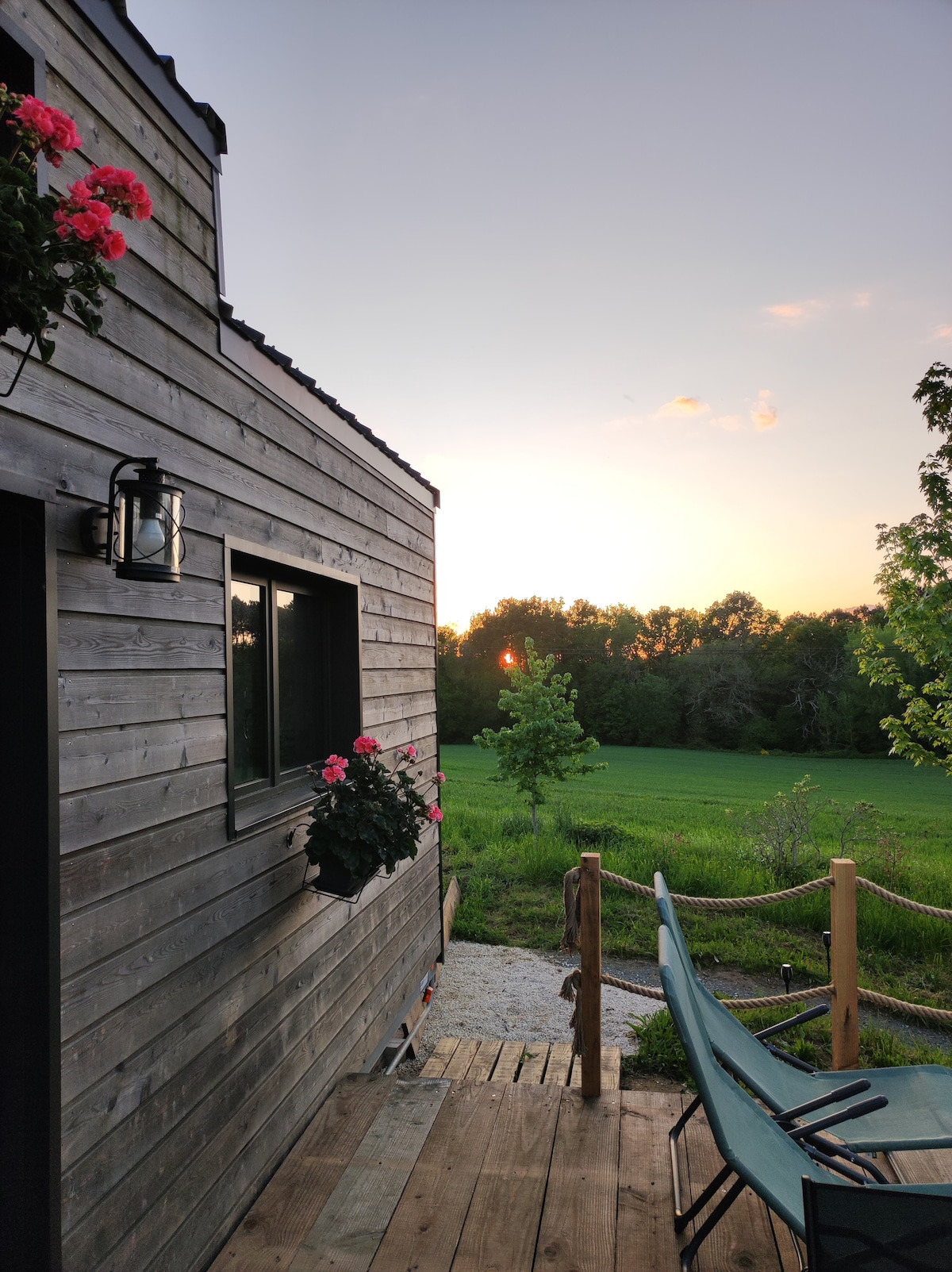 A wooden deck leads to a tiny house, framed by lush greenery and blooming pink flowers in window boxes. The sunset casts a warm glow over the expansive field in the background, creating a serene atmosphere.