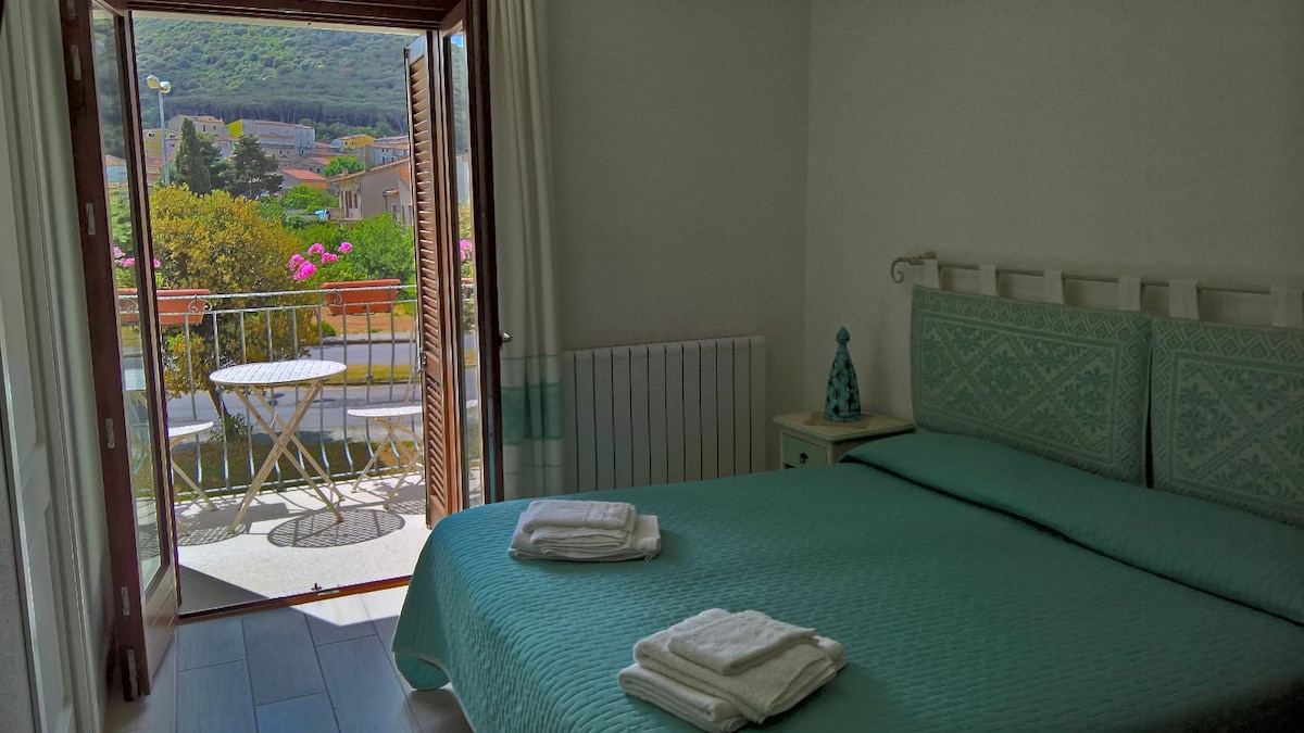 A comfortable bedroom is featured with a spacious bed covered in a light turquoise quilt. Natural light enters through the open double doors, leading to a small balcony with a table and two chairs, overlooking a garden with blooming flowers.
