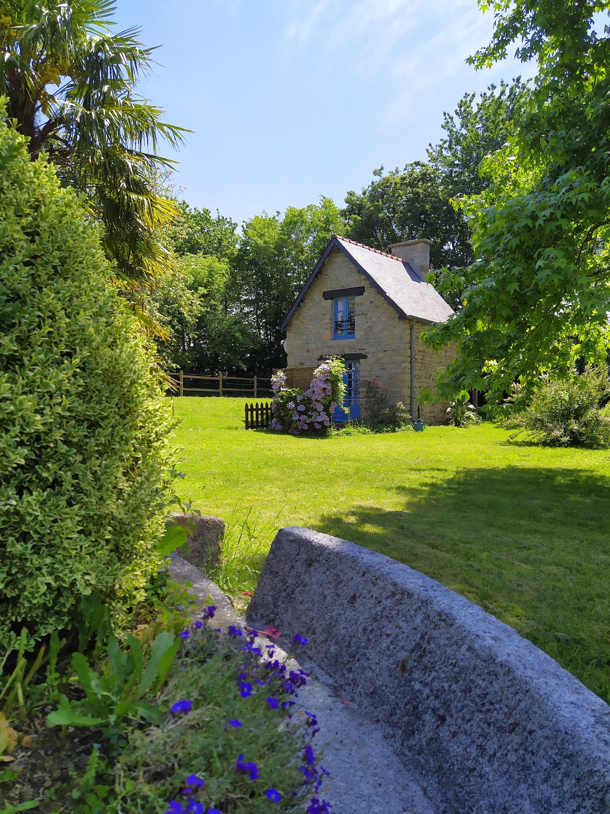 A charming independent house is surrounded by lush greenery and flowering plants. The structure features a sloped roof and vibrant blue window frames, set against a serene landscape that invites relaxation. A grassy area leads to a wooden fence, enhancing the tranquil rural setting.