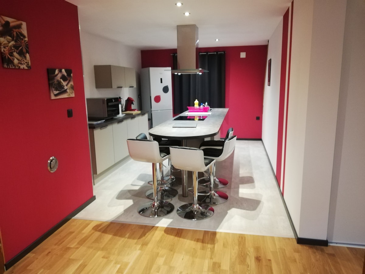 A modern kitchen space showcases a central island with high stools arranged around it. Sleek cabinetry, a refrigerator, and essential appliances are visible against vibrant red accent walls. Natural light brightens the area, highlighting the clean lines and contemporary design.