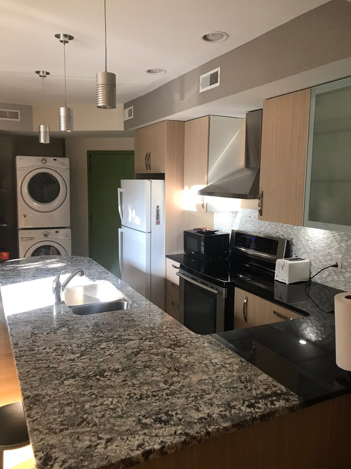 A modern kitchen features sleek, light wood cabinetry and a granite countertop. Stainless steel appliances include a refrigerator, microwave, and oven. In the corner, a stacked washer and dryer are visible. Soft lighting illuminates the workspace, creating a functional and inviting cooking area.
