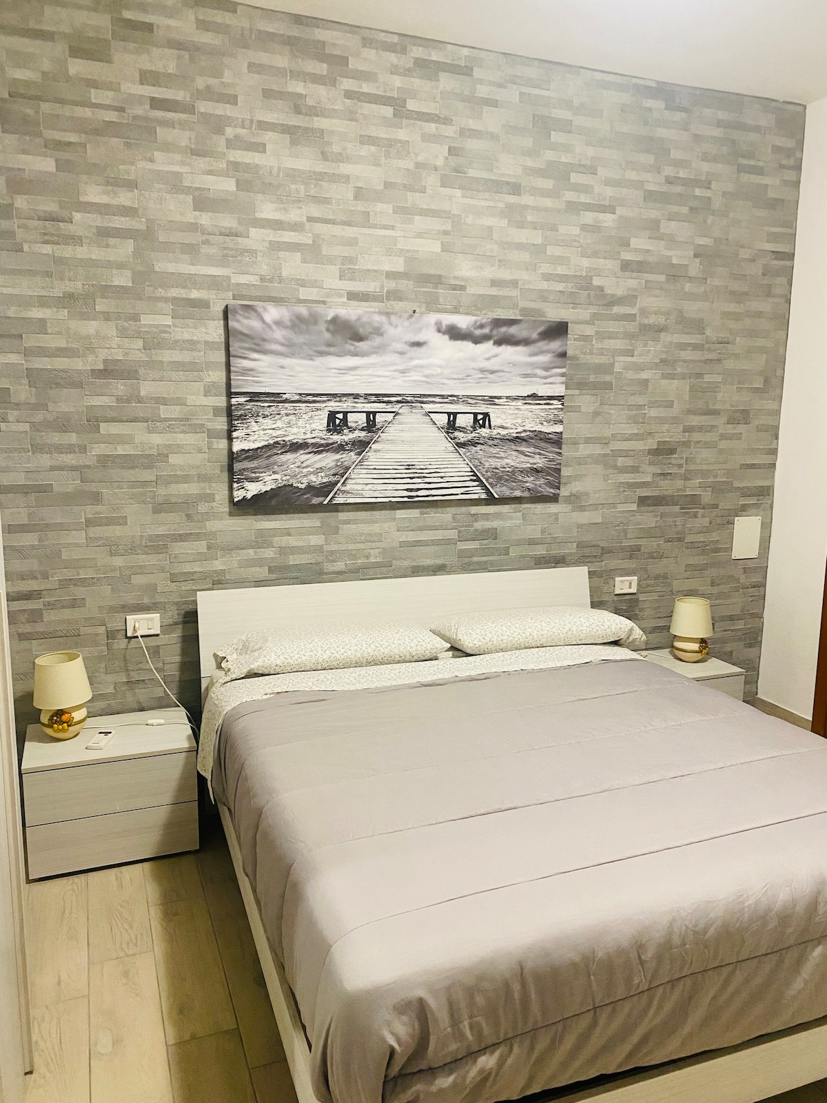 A cozy sleeping area is presented with light-colored walls and a textured gray accent wall. The spacious bed, dressed in neutral linens, is complemented by bedside tables that hold elegant lamps. A black and white art piece depicting a pier adds a tranquil touch to the room.